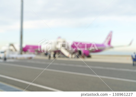 Blurred shot of a pink and white commercial airplane parked on an airport tarmac during the day. Passenger stairs are attached for boarding. Abstract travel concept with soft focus background. Blurred shot of a pink and white commercial airplane parked on an airport tarmac during the day. Passenger stairs are attached for boarding. Abstract travel concept with soft focus background. 138961227