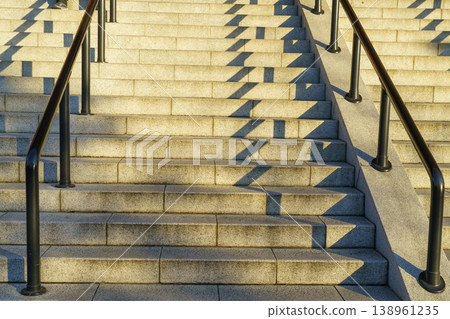 Wide view of concrete outdoor stairs with black handrails and dramatic geometric shadows under bright sunlight. Architectural detail of stone steps in a modern city during golden hour. 138961235