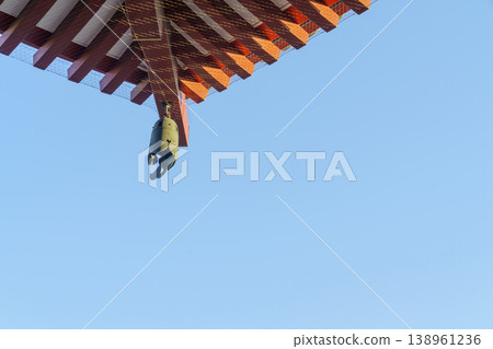 Traditional Japanese temple roof corner with a bronze hanging bell. Red wooden eaves against a clear blue sky. Cultural architecture detail showing Zen aesthetic and historical design. 138961236