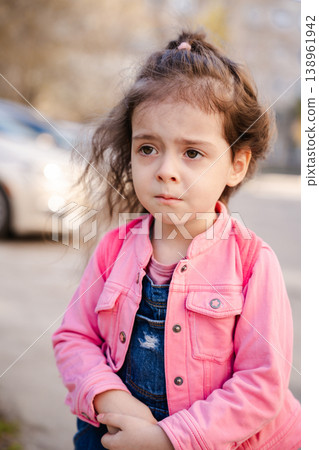 Little girl with curly brown hair wearing pink jacket standing outdoors near parked car and looking concerned on street. Childhood, emotion, urban lifestyle concept Little girl with curly brown hair wearing pink jacket standing outdoors near parked car and looking concerned on street. Childhood, emotion, urban lifestyle concept 138961942