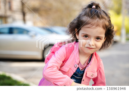 Little girl with brown hair wearing pink jacket and backpack leaning forward outdoors near parked car in residential street. Childhood, school, lifestyle, urban daily life concept Little girl with brown hair wearing pink jacket and backpack leaning forward outdoors near parked car in residential street. Childhood, school, lifestyle, urban daily life concept 138961946