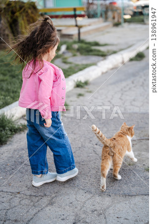 Little girl walking on street following cat with curious interested posture in residential neighborhood outdoors. Concept of childhood exploration animals and urban lifestyle Little girl walking on street following cat with curious interested posture in residential neighborhood outdoors. Concept of childhood exploration animals and urban lifestyle 138961947