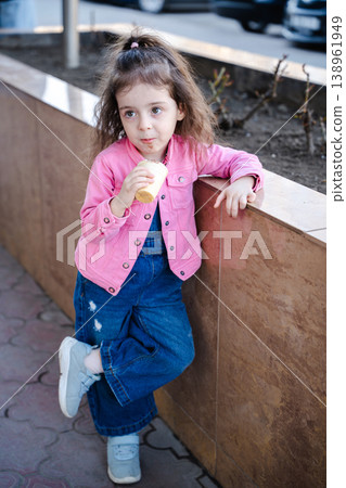 Little girl eating ice cream cone while leaning on urban planter outdoors and looking aside with thoughtful expression. Concept of childhood snack and casual city lifestyle Little girl eating ice cream cone while leaning on urban planter outdoors and looking aside with thoughtful expression. Concept of childhood snack and casual city lifestyle 138961949