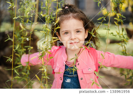 Little girl standing among green branches holding them with hands and looking at camera with calm curious expression outdoors. Concept of childhood nature exploration emotion and lifestyle Little girl standing among green branches holding them with hands and looking at camera with calm curious expression outdoors. Concept of childhood nature exploration emotion and lifestyle 138961956