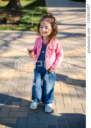 Little girl walking on paved path in park holding ice cream cone and looking aside with calm satisfied expression. Concept of childhood walk and outdoor leisure Little girl walking on paved path in park holding ice cream cone and looking aside with calm satisfied expression. Concept of childhood walk and outdoor leisure 138961957