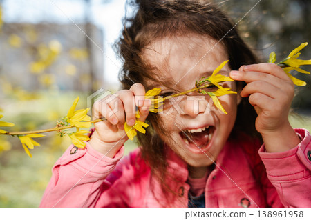 Little girl holding yellow flowering branch in front of face laughing with playful excited expression outdoors in sunlight. Concept of childhood emotion joy play and nature Little girl holding yellow flowering branch in front of face laughing with playful excited expression outdoors in sunlight. Concept of childhood emotion joy play and nature 138961958