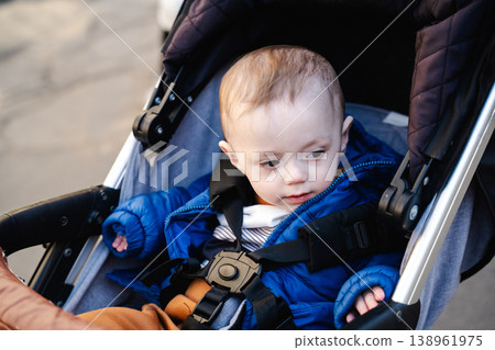 Infant child sitting in a stroller secured with safety harness wearing blue jacket outdoors on a walk in urban area. Concept of childcare parenting safety transport and daily life Infant child sitting in a stroller secured with safety harness wearing blue jacket outdoors on a walk in urban area. Concept of childcare parenting safety transport and daily life 138961975
