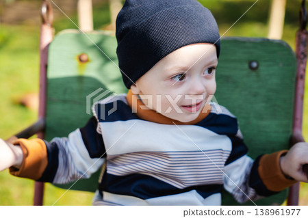 Infant child sitting on playground swing seat wearing black hat and striped sweater looking aside with curious expression outdoors. Concept of childhood emotion leisure and play 138961977