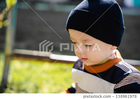 Close up portrait of infant child wearing black hat looking down with calm thoughtful expression outdoors in sunlight. Concept of childhood emotion observation and lifestyle 138961978