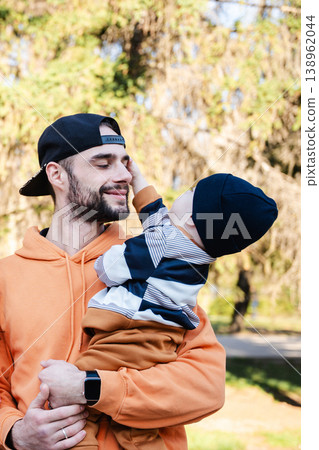 Father holding baby boy in arms outdoors while child touches his face and both show warm affectionate expressions. Concept of parenting and family bonding Father holding baby boy in arms outdoors while child touches his face and both show warm affectionate expressions. Concept of parenting and family bonding 138962044
