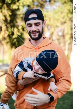 Father holding toddler boy horizontally in arms outdoors in park while child smiles and reaches forward. Concept of parenting and family bonding 138962047