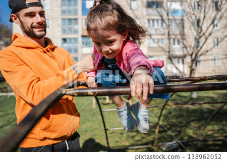 Little girl climbing on playground structure while father supports her from behind in outdoor park near residential buildings. Concept of childhood activity parenting support and trust Little girl climbing on playground structure while father supports her from behind in outdoor park near residential buildings. Concept of childhood activity parenting support and trust 138962052