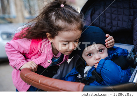 Little girl leaning over stroller touching baby head while infant sits inside looking at camera outdoors in city. Concept of sibling care family bonding and childhood 138962053