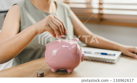 Woman putting coin into pink piggy bank on wooden table at home. Concept of financial planning, money saving for future, retirement investment, and personal budget management with copy space Woman putting coin into pink piggy bank on wooden table at home. Concept of financial planning, money saving for future, retirement investment, and personal budget management with copy space 138962617