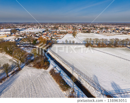 Aerial view of a snowy European town and countryside under a clear blue sky. Winter landscape with snow-covered fields, a narrow river canal, residential houses, and long shadows from the sun. Aerial view of a snowy European town and countryside under a clear blue sky. Winter landscape with snow-covered fields, a narrow river canal, residential houses, and long shadows from the sun. 138963051