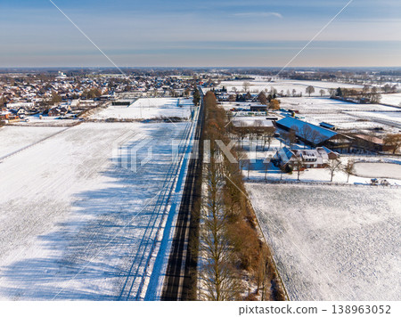 High-angle aerial view of a straight road passing through a snowy winter landscape in Europe. Featuring snow-covered fields, a small town, farm buildings, and long tree shadows on a sunny day. High-angle aerial view of a straight road passing through a snowy winter landscape in Europe. Featuring snow-covered fields, a small town, farm buildings, and long tree shadows on a sunny day. 138963052