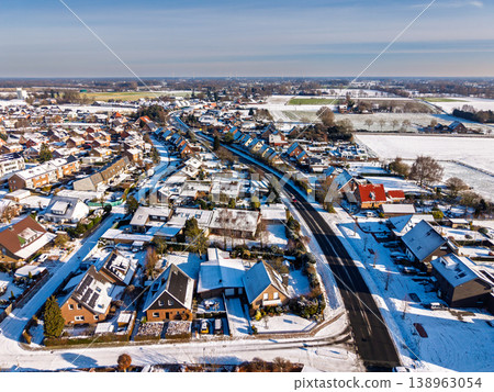 Aerial view of a quiet European town after a snowfall. The image shows snow-covered rooftops, clean asphalt roads, and sunny fields stretching to the horizon on a bright, cold winter morning. Aerial view of a quiet European town after a snowfall. The image shows snow-covered rooftops, clean asphalt roads, and sunny fields stretching to the horizon on a bright, cold winter morning. 138963054