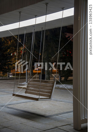 Wooden swing bench hanging on chains in a modern urban space at night, illuminated by soft warm lighting. 138963425