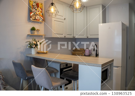 Modern compact kitchen in a studio apartment with a wooden bar counter, white cabinets, and warm pendant lighting, creating a cozy and stylish atmosphere. Modern compact kitchen in a studio apartment with a wooden bar counter, white cabinets, and warm pendant lighting, creating a cozy and stylish atmosphere. 138963525
