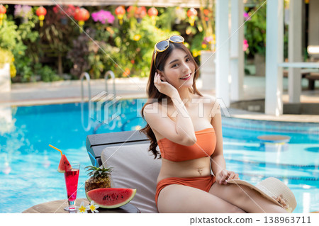 Sexy young asian woman in swimsuits in summer. beautiful woman in orange bikini with tropical drink sitting on sofa by swimming pool. Sexy young asian woman in swimsuits in summer. beautiful woman in orange bikini with tropical drink sitting on sofa by swimming pool. 138963711