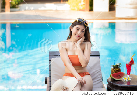 Sexy young asian woman in swimsuits in summer. beautiful woman in orange bikini with tropical drink sitting on sofa by swimming pool. 138963716