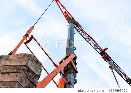 Low angle view of a construction crane with rigging and cables above stacked concrete blocks against a cloudy sky. Industrial building and heavy lifting Low angle view of a construction crane with rigging and cables above stacked concrete blocks against a cloudy sky. Industrial building and heavy lifting 138964411