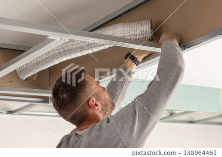 Construction worker handling metal profile frame in suspended ceiling system near flexible air duct 138964461