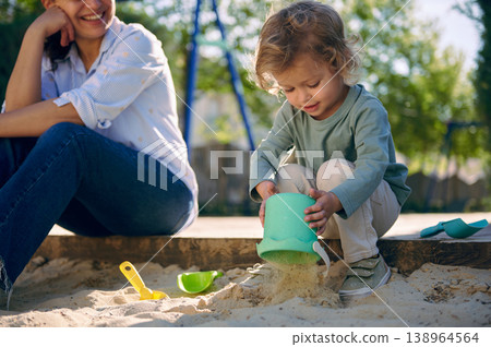 Mother Watching Toddler Play With Sand Bucket in Sunny Backyard Sandbox Mother Watching Toddler Play With Sand Bucket in Sunny Backyard Sandbox 138964564