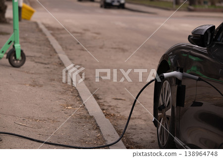 Electric Car Charging on City Street with Green Scooter in Blurred Background Electric Car Charging on City Street with Green Scooter in Blurred Background 138964748