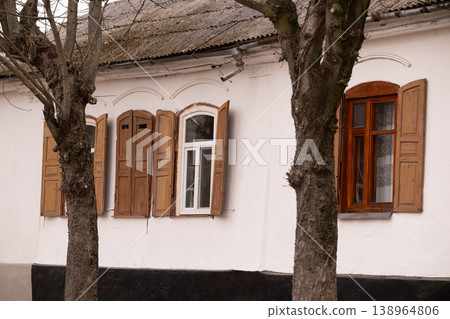 Old White House Facade with Wooden Shutters and Window Under Large Trees in Rustic Village Style Old White House Facade with Wooden Shutters and Window Under Large Trees in Rustic Village Style 138964806