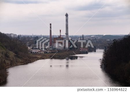 Industrial Factory with Tall Chimneys and Smoke Stacks overlooking Calm River under Cloudy Sky Industrial Factory with Tall Chimneys and Smoke Stacks overlooking Calm River under Cloudy Sky 138964822