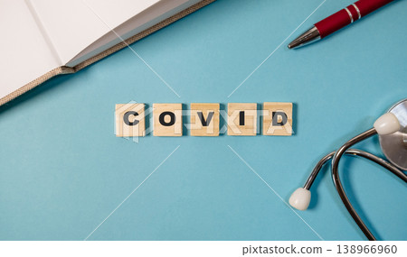 Wooden letter tiles spelling COVID on a blue background, accompanied by a stethoscope and pen, representing health awareness and the impact of illness on society 138966960