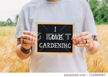 Individual holding a wooden-framed chalkboard sign that reads "I Love Gardening" in a golden field, surrounded by lush greenery and a serene atmosphere, expressing passion for gardening Individual holding a wooden-framed chalkboard sign that reads "I Love Gardening" in a golden field, surrounded by lush greenery and a serene atmosphere, expressing passion for gardening 138966967