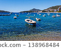 Small boats anchored in crystal clear turquoise water near Adriatic coast with coastal town and mountains in background, sunny summer day, peaceful seaside landscape 138968484