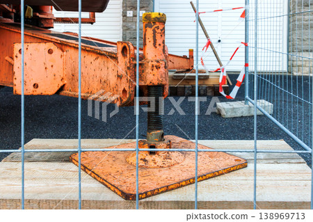 Close up of a rusty crane outrigger support foot on a metal base plate through a temporary safety fence at a roadwork construction site. 138969713