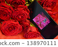 On the smartphone screen is a photograph of a rose lying among real red roses. 138970111