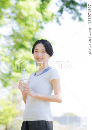 一位女性在戶外綠樹環繞中補充水分的側面肖像，她可能是在散步或跑步時。 138971867