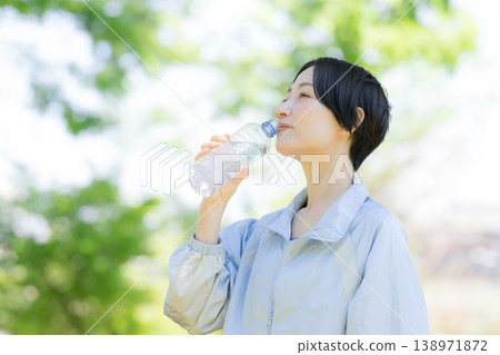 一位女性在戶外綠樹環繞中補充水分的側面肖像；她在運動或鍛煉間隙喝瓶裝水。 138971872