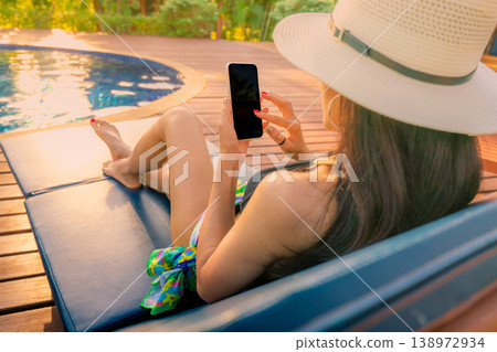 Asian woman wearing hat and swimsuit sitting on chair by swimming pool using smartphone during summer vacation, luxury lifestyle concept with manicure detail, checking news online in tropical resort. 138972934