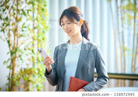 一位年輕的職業婦女在辦公室裡一邊使用智慧型手機一邊微笑。 一位年輕的職業婦女在辦公室裡一邊使用智慧型手機一邊微笑。 138972995