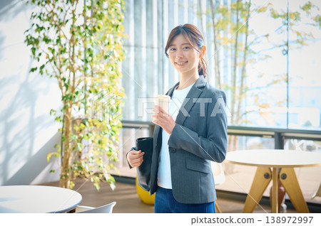 一位女商人站在辦公室裡，面帶微笑，手裡拿著一杯咖啡。 138972997