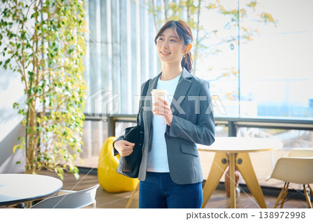 The lifestyle of a businesswoman standing with a smile, holding a coffee in the bright office cafe space. The lifestyle of a businesswoman standing with a smile, holding a coffee in the bright office cafe space. 138972998