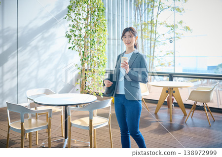 A businesswoman smiles relaxed, holding a cup of coffee in a brightly lit office cafe. A businesswoman smiles relaxed, holding a cup of coffee in a brightly lit office cafe. 138972999