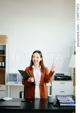 Asian woman, smiling and working at her desk in an office environment. She is wearing a business suit and holding documents while using a laptop. Asian woman, smiling and working at her desk in an office environment. She is wearing a business suit and holding documents while using a laptop. 138973973