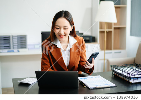 Asian businesswoman in brown blazer working on laptop, analyzing financial data with smartphone. at office Asian businesswoman in brown blazer working on laptop, analyzing financial data with smartphone. at office 138974010