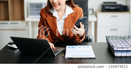 Asian woman, smiling and working at her desk in an office environment. She is wearing a business suit and holding documents while using a laptop. 138974012