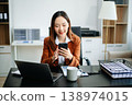 Asian woman, smiling and working at her desk in an office environment. She is wearing a business suit and holding documents while using a laptop. 138974015
