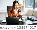 Asian businesswoman in brown blazer working on laptop, analyzing financial data with smartphone. at office 138974017