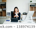 Asian woman, smiling and working at her desk in an office environment. She is wearing a business suit and holding documents while using a laptop. 138974139