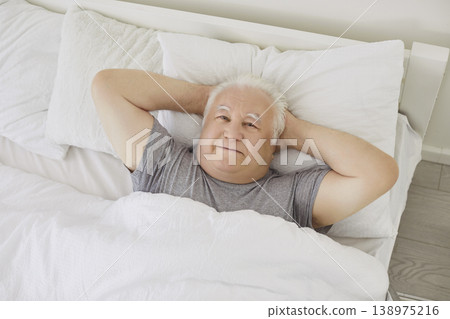 Happy smiling retired senior man lying with hands behind head in bed in the morning Happy smiling retired senior man lying with hands behind head in bed in the morning 138975216